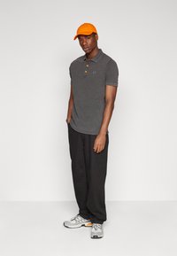 Polo gris con cuello en contraste, tapeta de tres botones y logo; combinado con pantalones negros y una gorra naranja; zapatos de running grises.