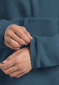Pullover in pile di colore blu scuro, con un tessuto testurizzato, polsini a costine e maniche leggermente arrotolate, che mostrano mani tatuate.