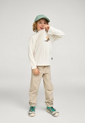 Child wearing beige pants, white long-sleeve shirt, and green cap, lifting shirt to mouth while standing on a plain background.