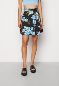 Woman wearing a black layered skirt with blue and white floral print, blue crop top, and black platform slide sandals standing against a white wall.