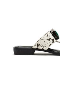 Sandales plates avec une base blanche présentant un motif à pois noirs, un accent en gemstone verte et une semelle en caoutchouc noir.