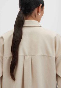 Donna con lunghi capelli scuri in una coda di cavallo bassa, che indossa un indumento beige con colletto, vista da dietro su uno sfondo neutro.