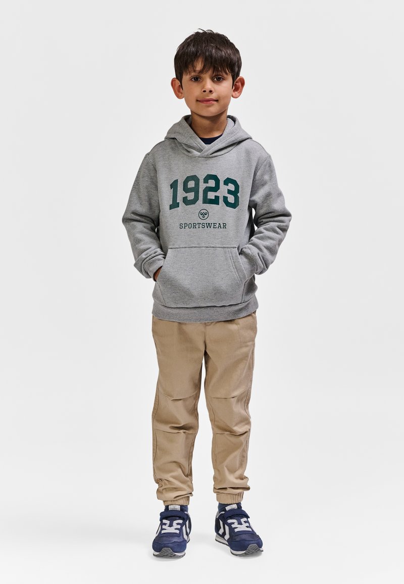 Grå hættetrøje med "1923 SPORTSWEAR" trykt i mørkegrøn, kombineret med lysebeige bukser og marineblå sko med hvide detaljer.