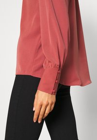 Langärmlige, hellrosa Bluse mit glatter Textur und Knopfdetails an den Ärmeln. Kombiniert mit schwarzen, eng anliegende Hosen.