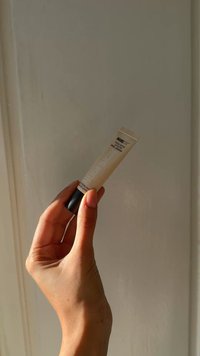 Nudestix NuDESKIN farget fuktighetskrem i squeeze tube, beige med svart hette. Hånd som holder tuben mot en lys veggbakgrunn.