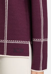 Burgundská pletená svetr s bílým geometrickým prošíváním. Obsahuje boční rozparek a logo "SCSY" na okraji, což zvýrazňuje strukturovaný vzhled.