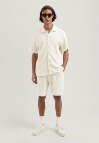 Rövidujjú, texturált ing és azonos színű, off-white rövidnadrág geometrikus mintával. Napszemüveggel és sportcipővel viselve.