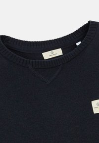 Pull en maille bleu marine avec col rond côtelé et étiquettes rectangulaires blanches de la marque au col et sur la poitrine portant la mention "Serge Blanco."