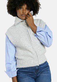 Pelēks adīts džemperis ar augstu apkakli un ar rāvējslēdzēju, uzvilkts virs zila un balta svītrainā garām piedurknēm krekla. Brīvs piegriezums, ērta tekstūra.