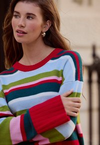 Multicolored striped sweater featuring red, green, pink, and blue hues. Ribbed red cuffs and neckline, soft texture, relaxed fit.