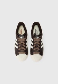 Para brązowych sneakersów Adidas z białymi noskami i trzema białymi paskami, widziana z góry na białym tle.