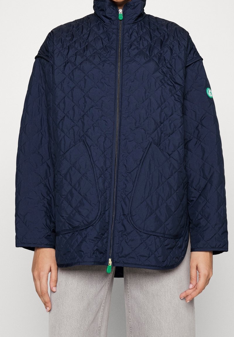 Chaqueta acolchada en azul marino con cuello alto, cierre frontal de cremallera y grandes bolsillos con parche. Presenta un tirador de cremallera verde y un acento de logo en la manga.