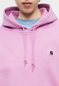 Άτομο που φοράει ανοιχτό μωβ φούτερ με κορδόνια και μικρό μαύρο λογότυπο στο στήθος, πάνω από ανοιχτό μπλε πουκάμισο.