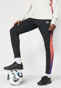 Pantalonii sport negri au dungi gradient de la portocaliu la mov pe laterale. Acestora le sunt asortați pantofii de fotbal negri și o minge cu un design pe fund.