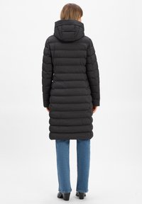 Schwarzer gepolsterter Mantel mit Kapuze, ausgestattet mit gestepptem Muster und vertikalen Nähten. Der Mantel hat ein mittellanges Design und wird mit blauen Jeans und schwarzen Schuhen kombiniert.