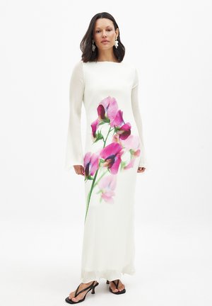 White ribbed long dress with bell sleeves, featuring a large pink flower print on the front. Worn with black strappy sandals.