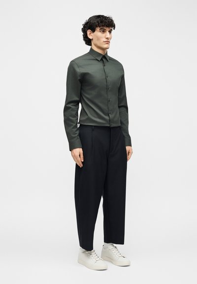 Άνδρας που στέκεται φορώντας σκούρο πράσινο πουκάμισο με κουμπιά, μαύρο παντελόνι με φαρδιές πιέτες και λευκά αθλητικά παπούτσια μπροστά σε απλό ανοιχτό φόντο.