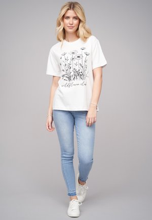 T-shirt en coton blanc avec un imprimé floral noir et le texte "wildflower club" en dessous. Assorti avec un jean skinny bleu clair et des baskets blanches.