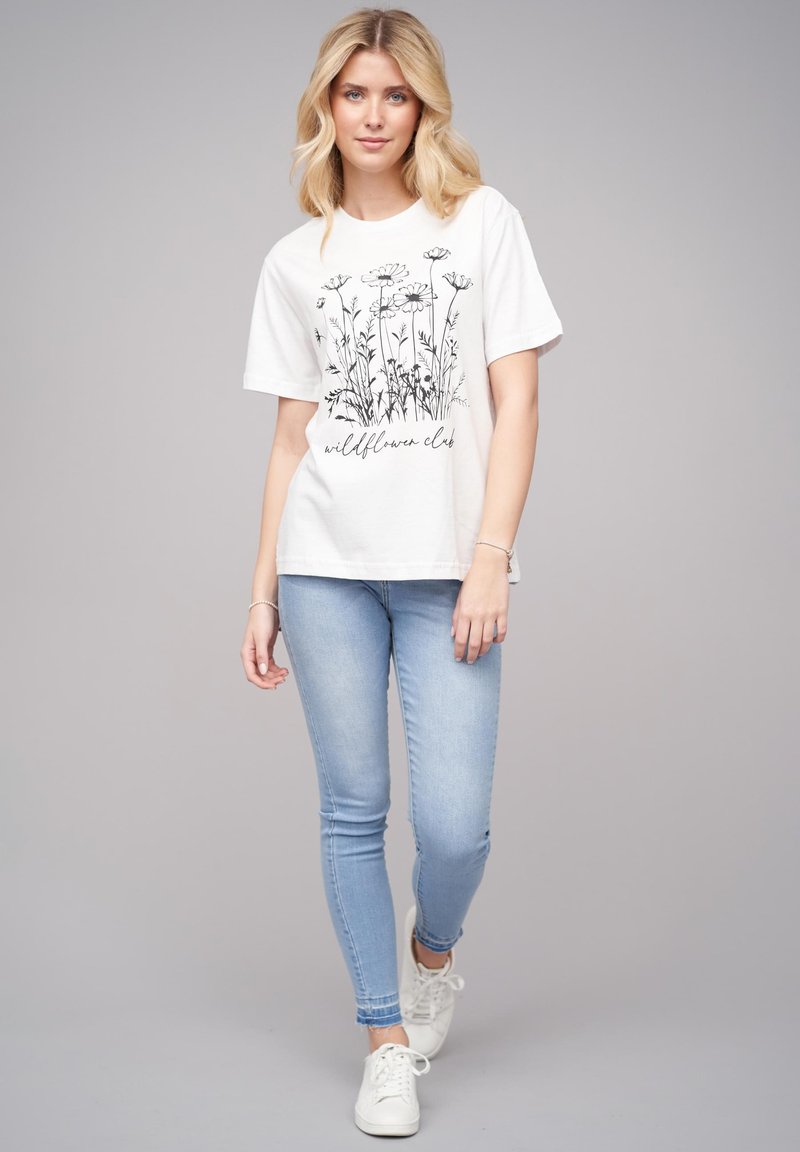 T-shirt en coton blanc avec un imprimé floral noir et le texte "wildflower club" en dessous. Assorti avec un jean skinny bleu clair et des baskets blanches.