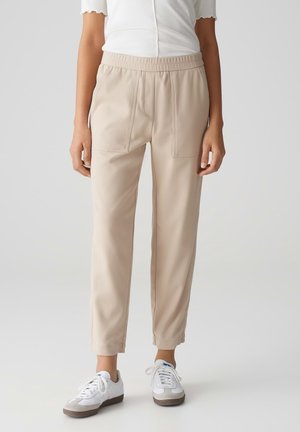 Kvinde iført beige ankellange bukser med elastisk talje og hvide sneakers, stående mod en ensfarvet lys baggrund.
