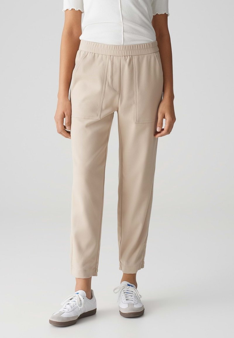 Femme portant un pantalon beige longueur cheville avec une taille élastique et des baskets blanches, debout devant un fond clair uni.