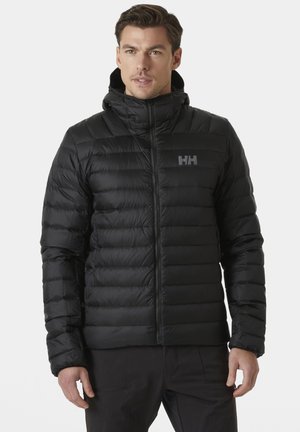 Man draagt een zwart doorgestikt gewatteerd jack met capuchon, een rits aan de voorkant en een merklogo op de borst, staand tegen een effen achtergrond.