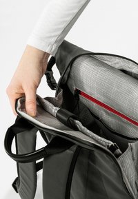 Zaino nero e resistente con rivestimento a scacchi grigi. Dotato di una tasca con zip rossa e vari scomparti. Maniglia per il trasporto nella parte superiore.
