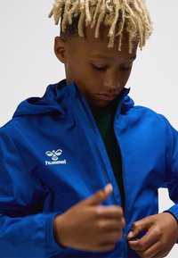 Blå lynlåstrøje med hætte. Fremstillet af glat, letvægtsmateriale. Har en hvid logo på brystet og elastiske ærmekanter.