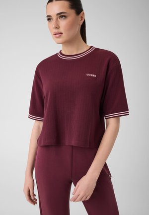 Kvinde iført en burgundy Guess kortærmet ribbet top med hvidstribet krave og ærmekanter, kombineret med matchende burgundy bukser.