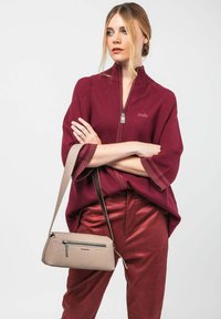 Blonde Frau mit einem Haarknoten trägt einen burgunderfarbenen Reißverschluss-Oberteil und Hose, hält eine beigen Schultertasche mit einer vorderen Reißverschlusstasche.