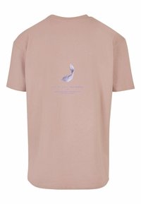 Světle růžové bavlněné tričko s grafickým motivem fialového peří a textem na zadní straně. Design zdůrazňuje jednoduchost a jemnost.