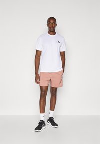Hvid trænings T-shirt med lille sort logo, parret med lyserøde shorts. Sorte træningssko og hvide sokker fuldender outfittet.