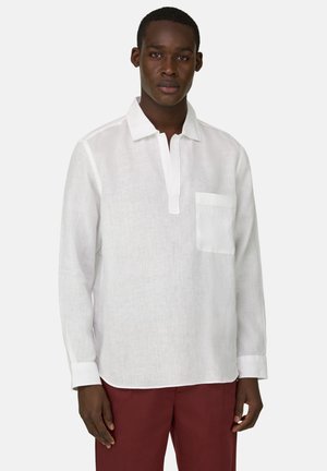 Homme portant une chemise en lin blanche à manches longues avec une poche poitrine et un pantalon bordeaux, debout devant un fond uni.