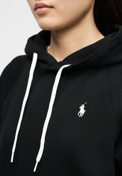 Κοντινό πλάνο ενός ατόμου που φοράει μαύρο φούτερ με κουκούλα, με λευκά κορδόνια και μικρό λευκό λογότυπο παίκτη πόλο στο στήθος.