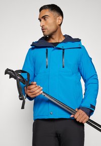 Blaue wasserdichte Jacke mit einem Reißverschluss auf der Vorderseite, zwei Brusttaschen und einer gefütterten Kapuze. Verfügt über verstellbare Ärmelbündchen und robusten Stoff. Hält einen Trekkingstock.