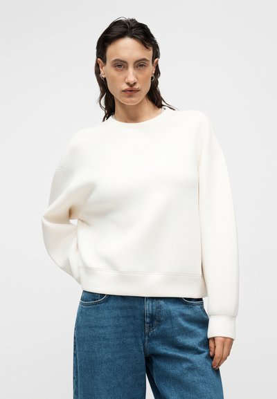 Φούτερ σε κρεμ απόχρωση με στρογγυλή λαιμόκοψη, φαρδιά εφαρμογή και ριμπ μανσέτες, συνδυασμένο με μπλε τζιν παντελόνι. Απλός, μινιμαλιστικός σχεδιασμός.