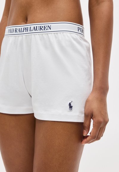 Άποψη από τη μέση έως το μέσο μηρού ενός ατόμου που φορά λευκά boxer Ralph Lauren Polo με λογότυπο στην ζώνη και μικρό μπλε λογότυπο παίκτη πόλο στο αριστερό πόδι.