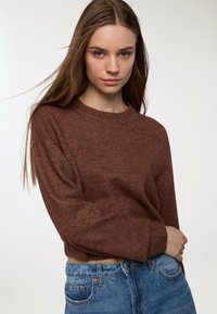 Giovane donna con lunghi capelli castani, che indossa un maglione di lana marrone e jeans blu, in piedi con un braccio incrociato sulla vita.