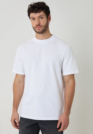Man met krullend donker haar, gekleed in een eenvoudige witte T-shirt met korte mouwen en een donkere broek, staand tegen een lichtgrijze achtergrond.