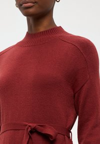 Μπορντώ πλεκτό πουλόβερ με ραμμένο λαιμό, μακριά μανίκια και λεπτομέρεια με δεσίματα στη μέση, που διαθέ�τει λεία υφή και χαλαρή εφαρμογή.