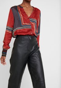 Blouse rouge à motif avec des accents bleus et crème, dotée d'un col en V et d'une fermeture à boutons, associée à un pantalon en cuir noir taille haute.