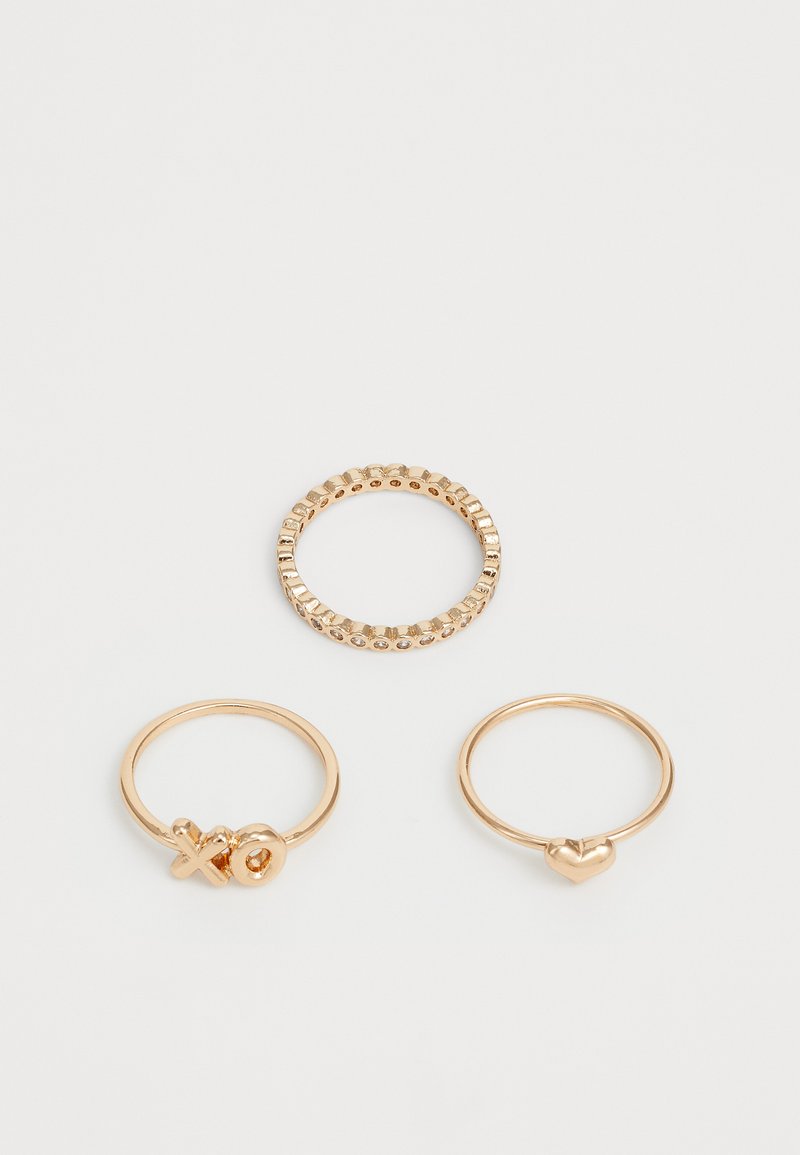 Tre anelli d'oro sottili, di cui uno con bordo a ondina, uno con charm XO e uno con charm a forma di cuore, su uno sfondo bianco semplice.