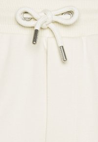 Joggers couleur crème avec une ceinture côtelée, dotés d'un cordon de serrage aux embouts métalliques. Le tissu semble doux avec une texture lisse.