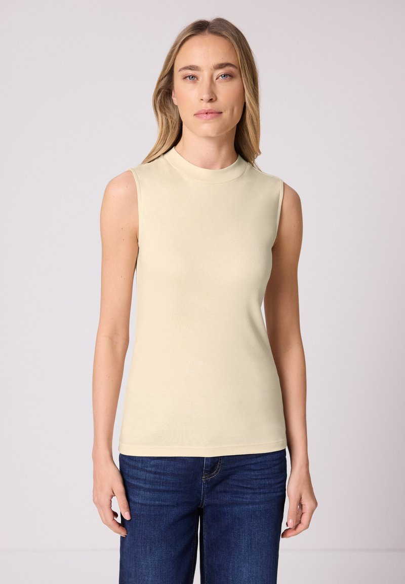 Femme aux longs cheveux blonds portant un haut beige sans manches et un jean bleu, debout contre un fond gris clair.