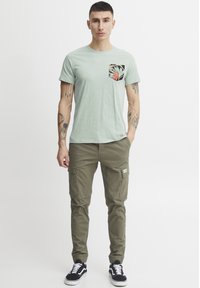 Camiseta de algodón verde claro con un bolsillo negro estampado, combinada con pantalones cargo oliva y zapatos negros con suelas blancas.
