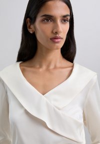 Blusa de seda en crema con un escote en V profundo y detalle de tela drapeada en el cuello. Textura suave y acentos de diseño minimalista.