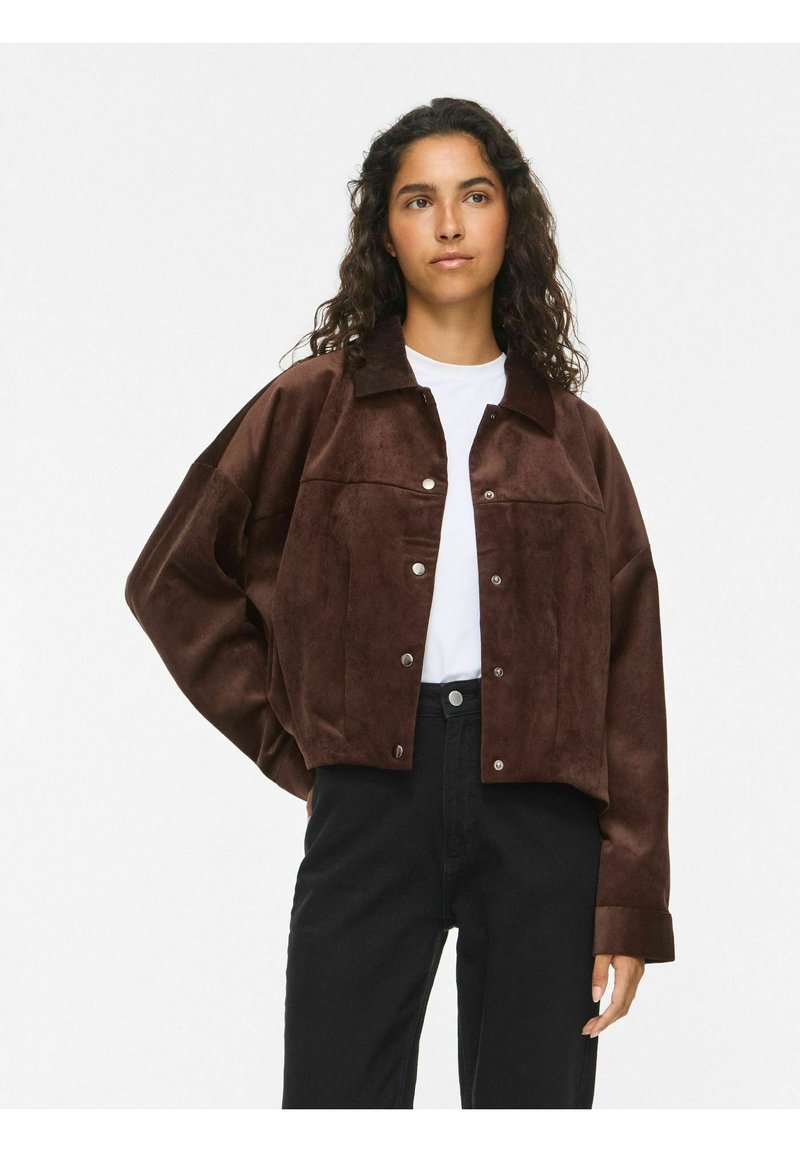 Femme aux cheveux bouclés et foncés porte une veste en daim marron par-dessus une chemise blanche et un pantalon noir, debout avec une main sur la hanche devant un fond uni.