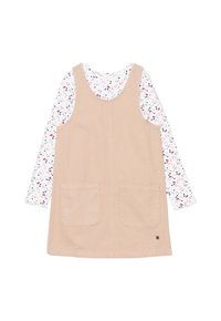 Beige Latzkleid mit zwei vorderen Taschen, über einem weißen Langarmshirt mit einem bunten Blumenmuster getragen.