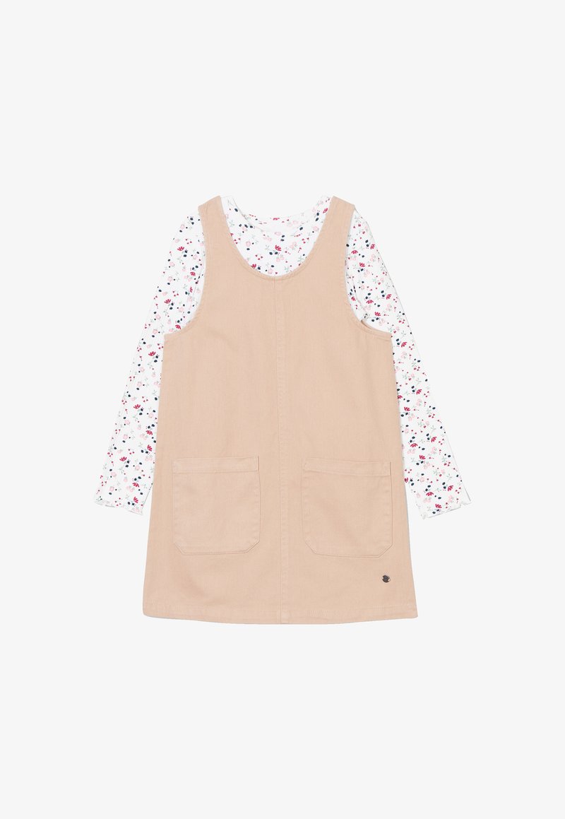 Abito salopette beige con due tasche frontali, indossato sopra una camicia bianca a maniche lunghe con un motivo floreale colorato.