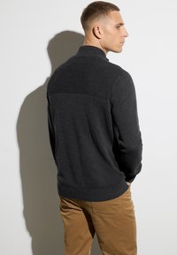 Mörkgrå stickad tröja med hög krage och ribbat mönster. Bärs med beige byxor som visar en avslappnad passform och en klassisk design.